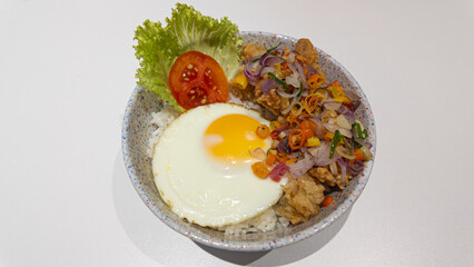 A fast food dish consisting of rice, fried egg, fried chicken in batter, sambal matah, lettuce, and tomato in a disposable bowl.
