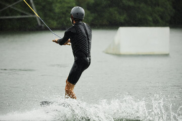 Back view of sportive adult man wearing wetsuit and practicing wakeboarding on lake waters moving towards ramps for action tricks copy space