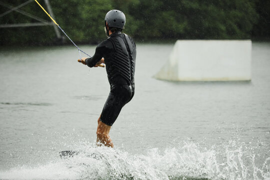 Back view of sportive adult man wearing wetsuit and practicing wakeboarding on lake waters moving towards ramps for action tricks copy space