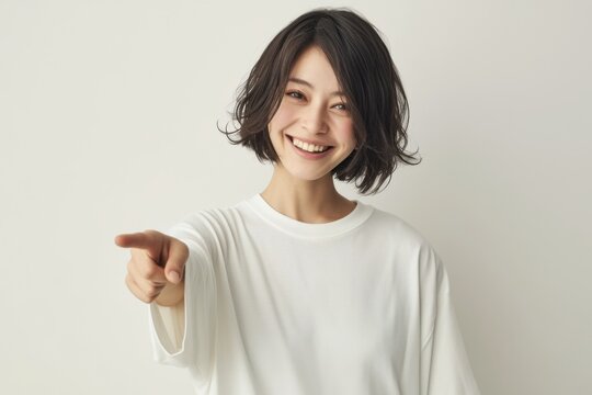 A cheerful Japanese woman in her early 20s smiles and points at the viewer with one hand, dressed in white against a plain background