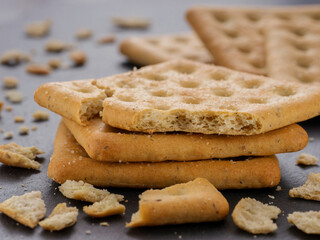 stack of square crackers with pieces and crumbs on slate gray background. Dry salt cracker cookies with fiber and dry spices