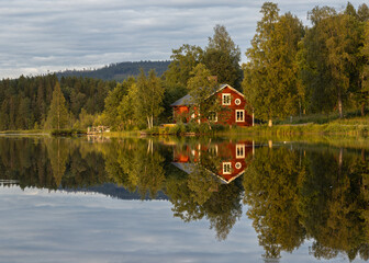 Swedish stuga by a calm lake on a cloudy summer day. The sunlight illuminates the green trees and...