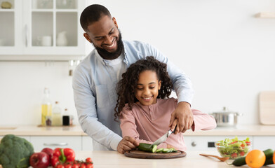 Bonding Concept. Cheerful african american father helping his daughter to cut vegetables, slicing cucumber with knife, teaching little girl how to prepare healthy fresh salad, cooking together at home