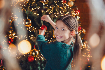 Photo of cute little girl decorate tree dressed green christmas outfit day light home party decoration interior room