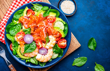 Healthy food salmon salad for keto diet with shrimp, avocado, spinach, cucumber, tomato, cashew, sesame seeds. Blue background, top view