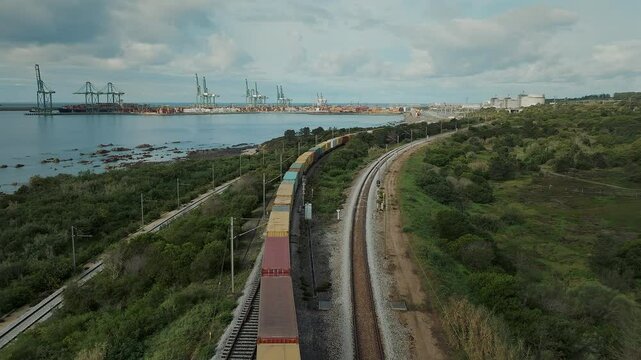 Aerial view of moving freight train with colorful wagons by the sea. Cargo train approaching the seaside port, delivering goods and industrial material. Transportation concept. 