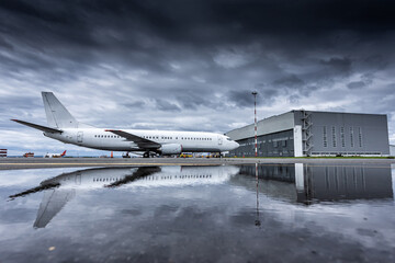 White passenger airplane at airport near aircraft hangar with reflection in puddle in cloudy weather