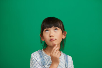 Fototapeta premium Asian child, girl and thinking in studio for future, education or remember by green background. Kid, ideas and problem solving for solution, choice or decision with development, process or memory