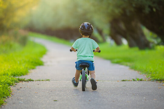 Little boy with helmet running and riding on first bike without pedals on sidewalk at city park in warm summer day. 2 years old toddler. Learning to keep balance. Back view.