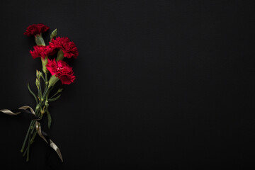 Fresh red carnation flowers with ribbon on dark black table background. Closeup. Condolence card. Empty place for emotional, sentimental text, quote or sayings. Top down view.