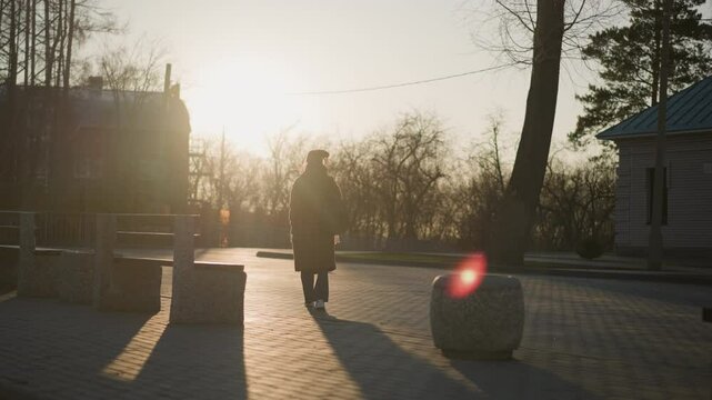 backshot of a woman wearing a brown coat, jeans, and white shoes, carrying a backpack as she walks alone through a park. The warm, golden light casts long shadows and creates a serene atmosphere