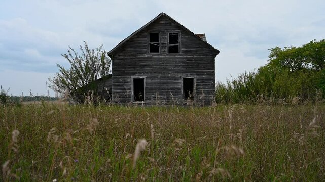 A dilapidated wooden farmhouse with missing windows is set in autumn colored long grass under a cloudy sky. The grass is swaying in a light wind.
