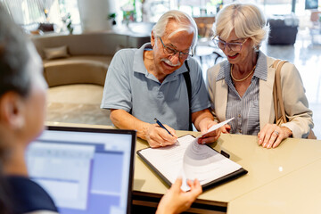 A smiling elderly couple stands in front of a hotel desk, filling out a check-in form or signing a...