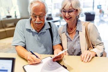 A smiling elderly couple stands in front of a hotel desk, filling out a check-in form or signing a...