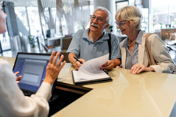 A smiling elderly couple stands in front of a hotel desk, filling out a check-in form or signing a...