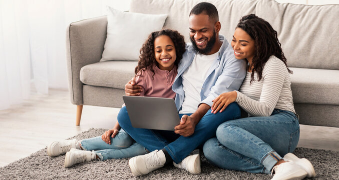 Family Weekend. Cheerful African American parents and their little daughter using laptop at home together, watching movie or browsing internet, sitting on the floor carpet in living room - Powered by Adobe