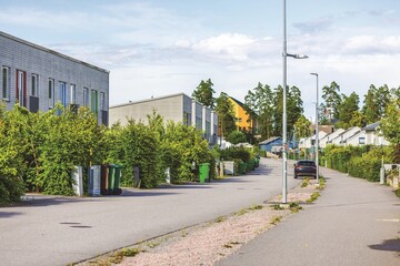 Residential street with modern townhouses, greenery, and trash bins along road under clouds sky. Sweden.