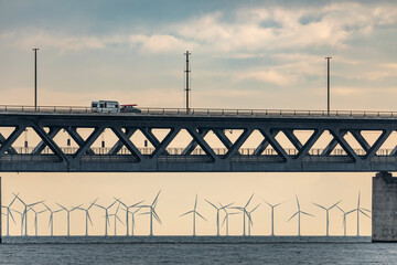 A caravan crosses an elevated bridge, next to a car with a surfboard on the roof. Below, the sea is home to a wind farm, with rows of turbines standing out on the horizon.
