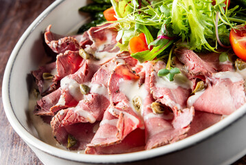 plate of sliced roast beef topped with sauce and capers, accompanied by fresh salad with mixed greens and cherry tomatoes. dish is elegantly presented on white plate, perfect for culinary photography.