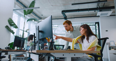 Attractive man helping his coworker solving work issues. Pretty woman sitting at desk with computer while manager standing behind. Female explaining to man something and pointing finger on screen.
