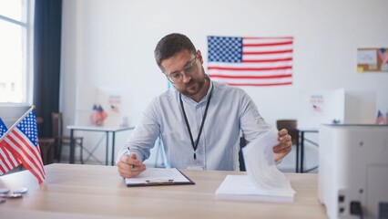 Handsome Caucasian male with beard sitting at desk in large office. Writing something with pen on...