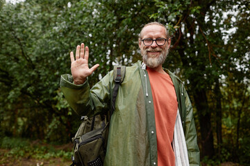 Waist up portrait of smiling senior man wearing raincoat waving at camera and enjoying tourism in nature, copy space