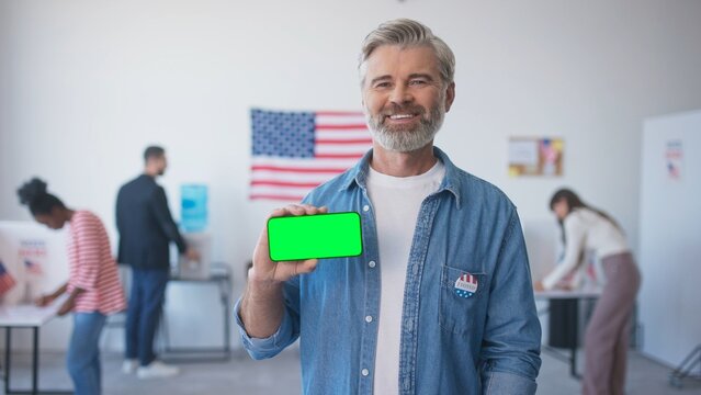 Man holding cell phone with chroma key in horizontal position. Voter showing to camera smartphone with green screen. Citizens of the United States of America voting in polling booth in background. - Powered by Adobe
