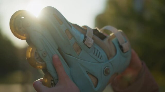 A close-up shot of a light blue rollerblade being held by a hand in a sunlit park. The sunlight filters through the background, creating a warm atmosphere, with the rollerblade prominently displayed