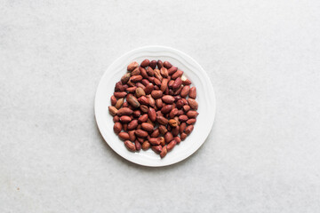 Overhead view of unpeeled raw peanuts on a white plate, flatlay of raw peanuts on a marble countertop, process of making roasted peanuts