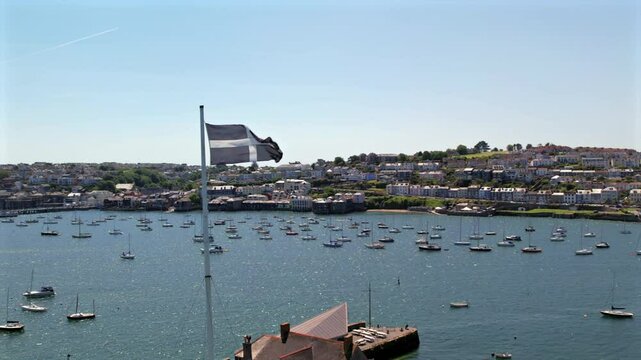 Aerial view of the Cornish flag waving over Falmouth harbor, with boats anchored and houses in the background on a clear day.