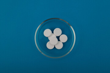 Chlorine tablets in glass Petri dish on blue background, top view. Chlorine tablets is used for disinfection of water, for bleaching fabrics, paper, wood. Cleaning product, bleach. Cl pills