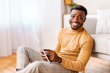 Positive Afro Man Using Tablet Computer Smiling To Camera Sitting On Floor At Home. Selective Focus