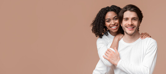 Romantic interracial couple embracing and posing together over yellow background, looking at...