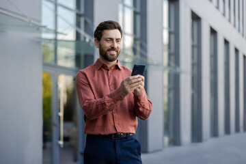 Mature male professional standing outside in city setting, holding phone. Man wearing casual attire, engaging with smartphone. Depicts business, communication, technology, and lifestyle concepts.