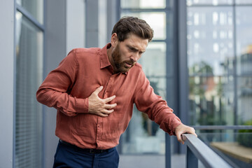 Adult male manager clutching chest in pain while standing on city street. Businessman shows signs...