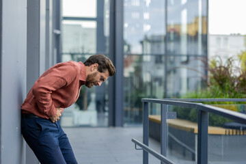 Man in city experiencing severe stomach pain. Male professional leaning against wall with pained...