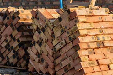 Piles of orange and brown bricks rest against a blurred backdrop at a construction site, suggesting preparation for upcoming building projects under bright sunlight