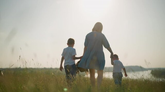 A back shot of a mother in a blue dress carrying a basket in one hand holding her younger son's hand with the other. Her older son walks beside them as they walk through a grassy field towards a lake