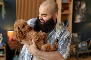 Smiling man gently cuddling fluffy brown dog in cozy living room surrounded by warm decor and personal items portrait of contentment and affection