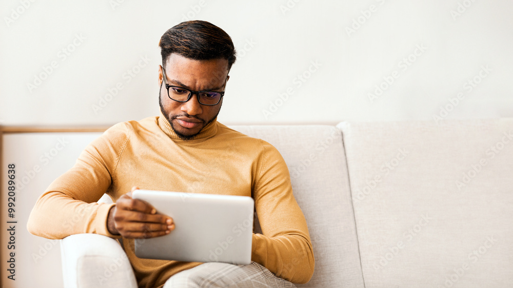 Poster serious afro entrepreneur man using digital tablet sitting on sofa at home. selective focus