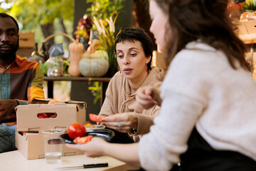 Female vendor sitting with multicultural couple, giving fresh fruits and veggies for tasting. Young customers enjoying a taste of natural homegrown organic slice of tomato at a farmers market.