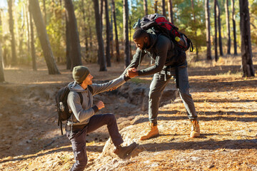 Two friends guys hiking together at countryside, black man helping caucasian guy to uphill, friendship concept