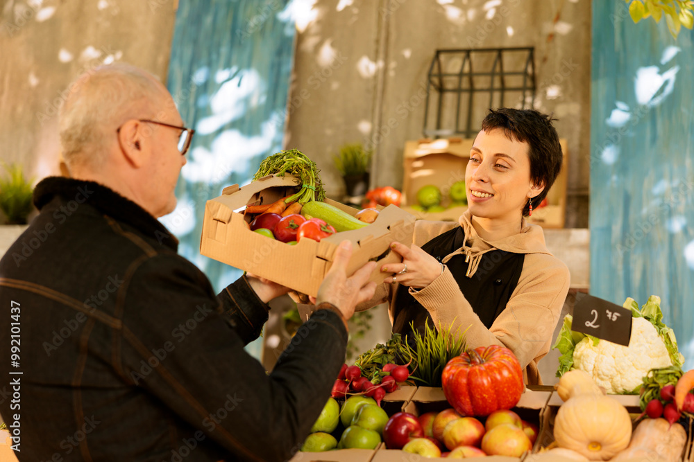 Wall mural Friendly female vendor giving box full of fresh organic fruits and vegetables to elderly customer while working at local farmers market. Farmer selling healthy locally grown farm food at harvest fair.