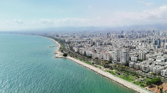 Coastline showing mersin city extending along turquoise water. Buildings and palm trees are visible. Mountains are seen in the distance
