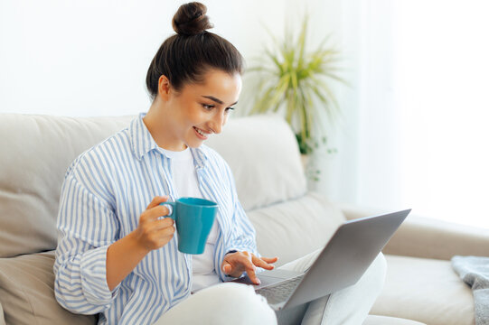 Positive indian or arabian woman, sits on a couch at home in a living room with a cup of tea or coffee, using laptop, working online, doing shopping, pays the bills, chatting in social media, smiles