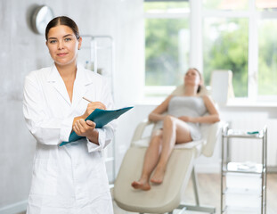 Obraz premium In beauty parlor of medical clinic female doctor stands with folder and fills out documents before manipulations. Girl patient is lying in chair in background and is waiting for procedure to begin