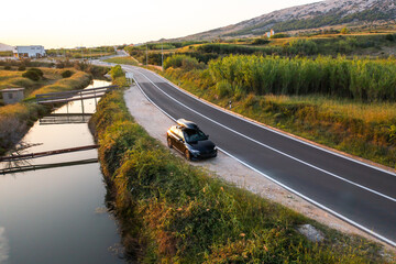 A black vehicle equipped with a roof rack is parked along a serene road, surrounded by greenery
