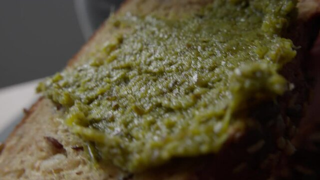 A knife spreads a generous layer of green pesto sauce over a slice of multigrain bread. The close-up captures the rich texture of the bread and the smooth, vibrant pesto.