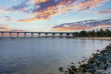 Fototapeta premium San Diego bay and the Coronado Bridge with ships sailing on blue ocean water, office buildings and skyscrapers along the coast at sunset at Coronado Tidelands Park in Coronado California