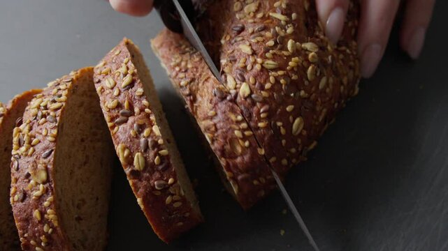 A top view shot of a person slicing a loaf of fresh multigrain bread with a knife. The bread is covered with seeds, showcasing a rustic texture and appetizing crust.